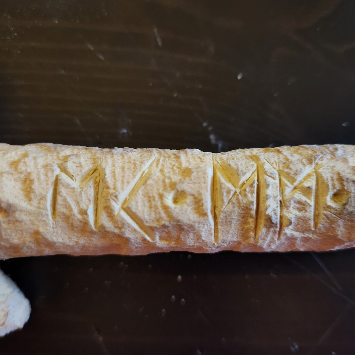 A section of pale stick with runes carved into the wood, highlighted by light and shadow.
