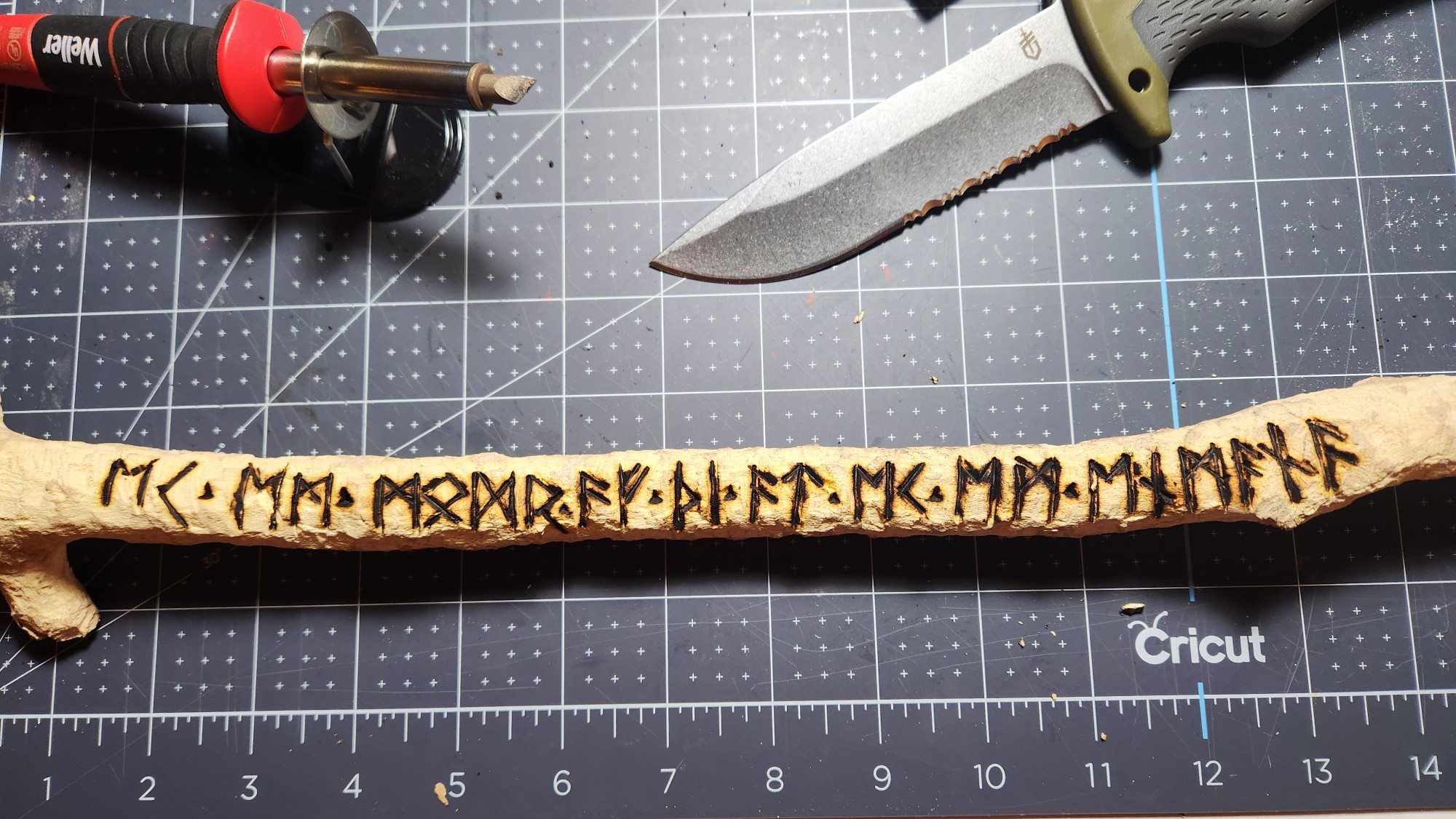 The completed woodburning stage, with thin runes at the start and thicker, charred runes toward the end. Also in the picture are the pyrograph and the outdoor utility knife used for carving, resting on a Cricut mat.