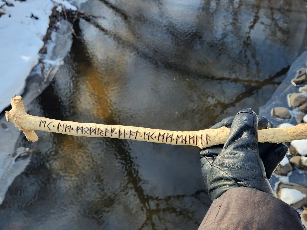 A gloved hand holds the rune stick over the rippled water of a creek. The banks are lined with snow-covered rocks.