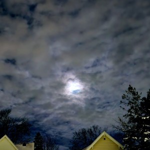 A glowing full moon behind stippled clouds, above a snowy neighborhood.