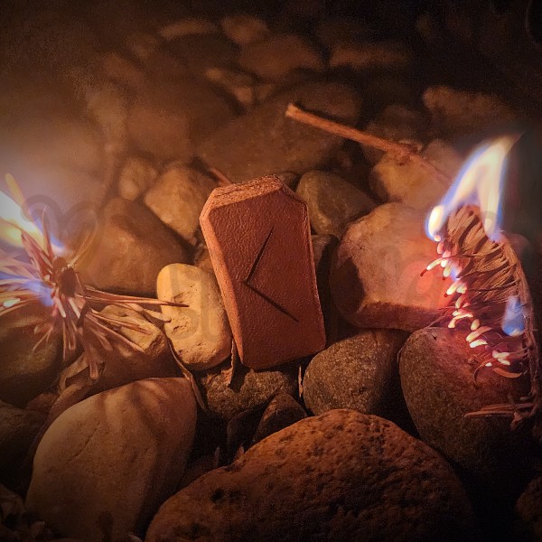 The rune Kenaz burned into a piece of leather, standing upright among stones. On either side a twig of old pine is burning.